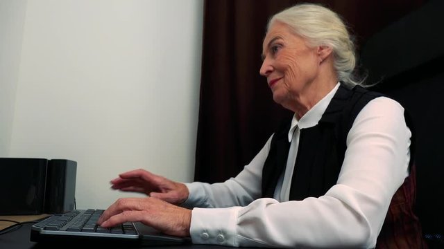 Old happy caucasian woman works on computer in home and smiles