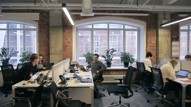 Young colleagues are chatting sitting at table with laptops in large company. Men talk actively, sitting on individual workplace while people are fussing indoors. Promising workteam is engaged in