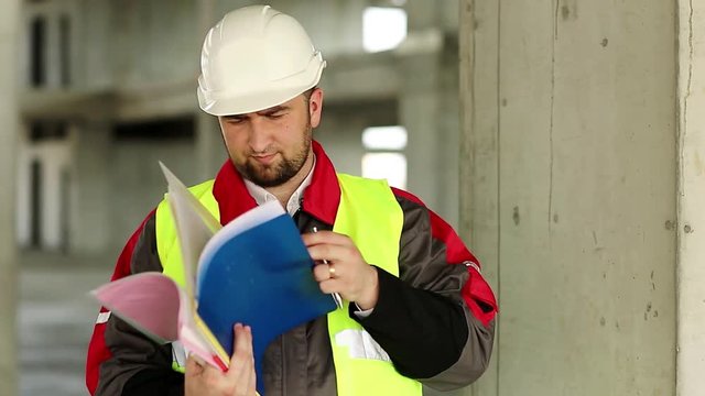 Building inspector with construction documentation at project site. Master builder in white hard hat with works papers at construction site