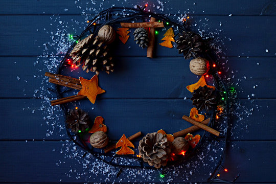 Christmas Wreath With Pine Cones, Garland Lights, Cinnamon, Walnuts, Snow And Dried Orange Decoration On Gray Wooden Background