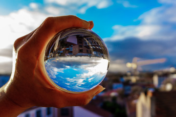 A hand holding a crystal ball for optical illusion. City as the background. Known as an orbuculum, is a crystal or glass ball and common fortune telling object. Performance of clairvoyance and scrying - obrazy, fototapety, plakaty