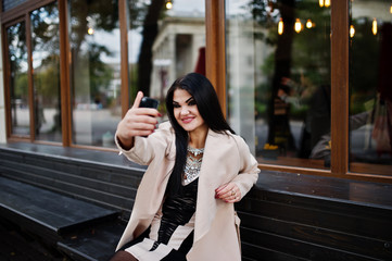 Black hair sexy woman in glasses and coat sitting on bench with mobile phone at hand and making selfie.