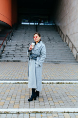 Vertical full body of casual woman posing on the street in cold windy day with cup of coffee. Outdoor photo of woman in trendy gray coat spending time in city and drinking latte.