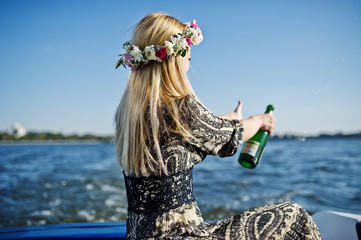 Blonde girl in wreath sitting on yacht at hen party and opening champagne.