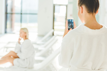 Obraz premium woman taking photo of her friend at spa center with smartphone