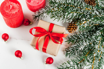 Christmas background for greeting card. Christmas tree branches with snow effect with festive red ribbon, pine cones, gift boxes and candles, on white marble background, copy space top view