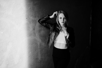 Elegant blonde girl wear on black leather jacket posing at streets of town background old wall with shadows.