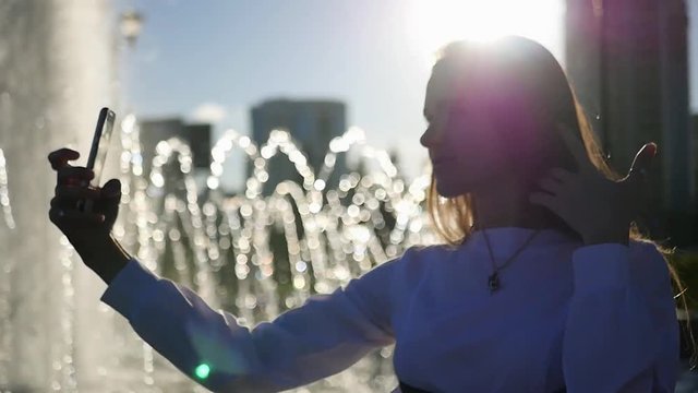A beautiful girl with long hair poses on the camera, takes a photo standing at the fountain with the blurred rays of the sun. HD, 1920x1080. slow motion.
