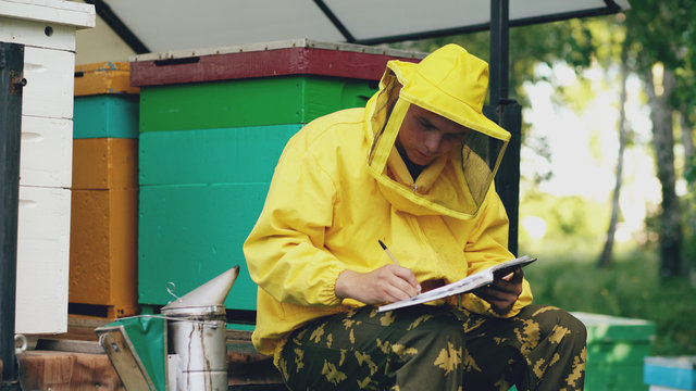 Young beekeeper man write in notepad checking harvest while sitting near beehives