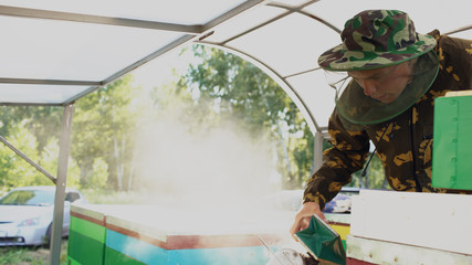 Young beekeeper man smoking bees away from beehive in apiary