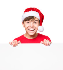 Teen boy wearing Santa Claus hat, posing behind white panel isolated on white background. Teenager child holding empty Christmas billboard. Young student peeping behind blank holidays board. 