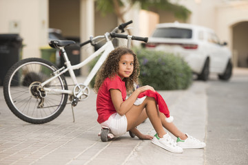 Fototapeta na wymiar A cute biracial girl sits and rest with her Santa hat on her knee on the side walk with her bicycle in the background