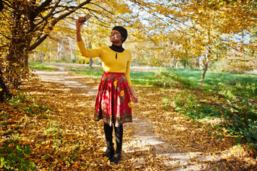 African american girl at yellow and red dress at golden autumn fall park making selfie on phone.