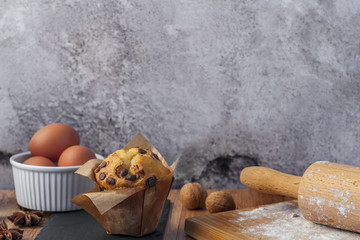Muffin with chocolate, eggs in a white bowl, cutting board, stone black plate, rolling pin, anise and walnuts on a wooden brown table against the background of an empty concrete wall