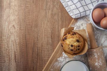 Muffin with milk chocolate, eggs in a white bowl, a glass of almond milk, a check cloth, a rolling pin and flour sprinkled on a brown wooden table with an empty place for text seen from the top