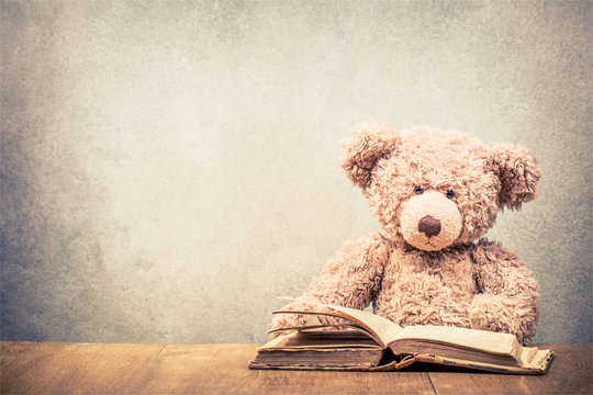 Retro Teddy Bear toy sitting at the old wooden desk with old book front concrete wall background. Vintage instagram style filtered photo