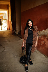 Fashion outdoor photo of gorgeous sensual woman with dark hair in elegant clothes and luxurious fur coat at old street with grunge walls.