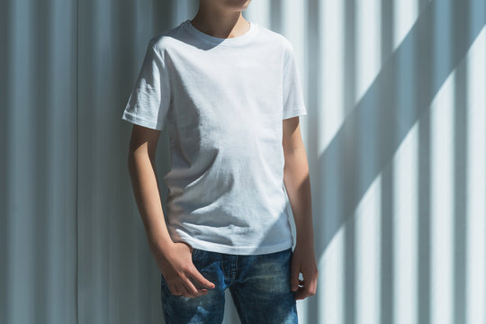 Front view. Young hipster boy dressed in white t-shirt is stands indoor against white wall. Mock up. Space for logo, text, image.