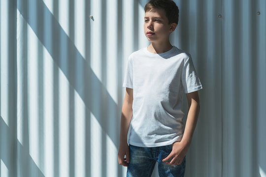 Front view. Young hipster boy dressed in white t-shirt is stands indoor against white wall. Mock up. Space for logo, text, image.