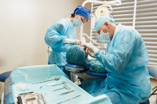 Male Old Professional Dentist Surgeon In Uniform And Female Assistant Helps To Performing Operation Install Dental Implant Teeth Of Woman Patient In Clinic Light Office With Modern Tools Equipment.