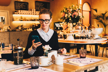 Young business woman in glasses sits in cafe at table, uses smartphone. On table is cup of coffee. Girl working, chatting, blogging, checking email. Online marketing, education, social media, network.