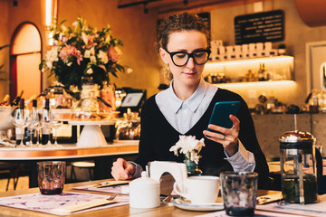 Young business woman in glasses sits in cafe at table, uses smartphone. On table is cup of coffee. Girl working, chatting, blogging, checking email. Online marketing, education, social media, network.