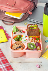 Lunch box with sandwiches, vegetable salad and fresh fruits and rice on the table near school backpack