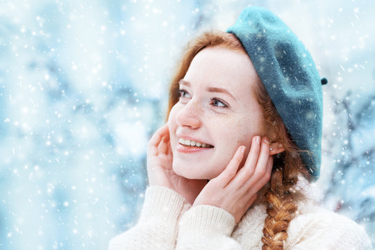 Happy smiling redhead girl with freckles touching her face, looking up. Model posing in street, wearing beret and sweater. Snowfall. Winter, Christmas holidays concept. Copy, empty space for text