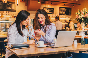 Two young happy women are sitting in cafe at table in front of laptop, using smartphone and laughing. On table paper notebook and cup of coffee. Girls are blogging, working, studying, learning online.
