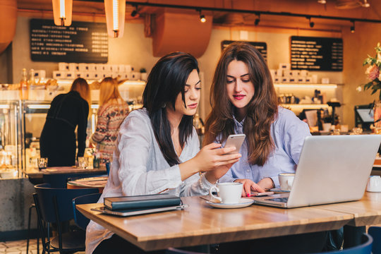 Two young happy women are sitting in cafe at table in front of laptop, using smartphone and laughing. On table paper notebook and cup of coffee. Girls are blogging, working, studying, learning online.