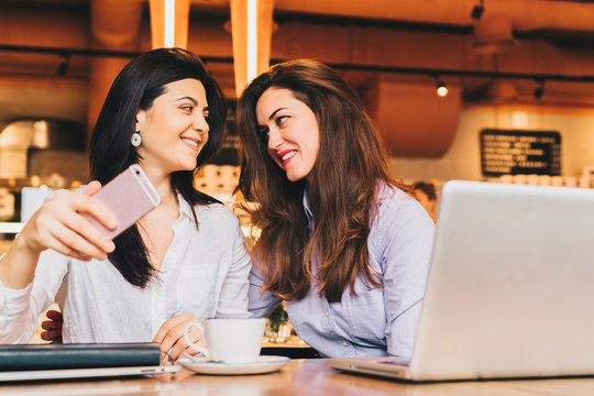 Two young happy women are sitting in cafe at table in front of laptop, using smartphone and smiling. On table paper notebook and cup of coffee. Girls are blogging, working, studying, learning online.