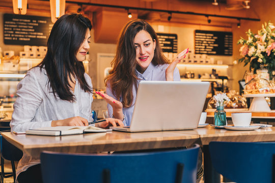 Two young businesswomen, bloggers, wearing in shirts are sitting in cafe at table and using laptop, working, blogging, online shopping. Girls look at monitor in amazement, delightedly, joyfully.