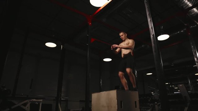Muscular man without a shirt performs vertical jumps on a wooden box. Aerobic exercise.