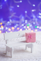 White little wooden bench with pink gift box on white knitted fabric on purple background with warm...
