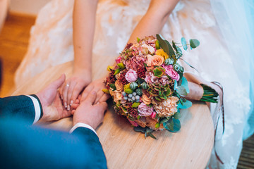 Hands of bride and groom with engagement rings on the table with wedding flowers bouquet.