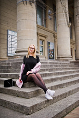 Fototapeta na wymiar Blonde girl at glasses and pink coat with handbag sitting on stairs of opera building.