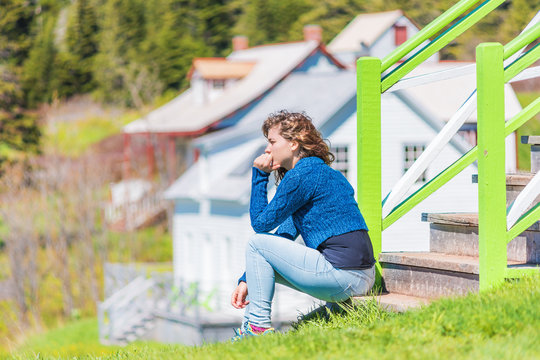 Young woman sitting on porch of colorful painted house in idyllic countryside summer scenery in Scandinavia or Canada with hand on face thinking