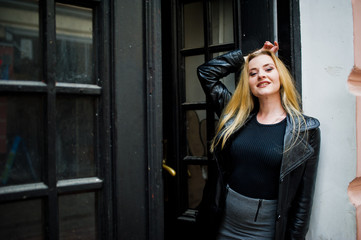 Fototapeta na wymiar Blonde fashionable girl in long black leather coat posed against wooden door at old house.