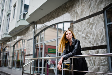 Naklejka premium Blonde fashionable girl in long black leather coat posed at iron railing.