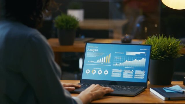 Female Analyst at Her Desk Works on a Laptop Showing Statistics, Graphs and Charts. She Works on the Wooden Table in Creative Office. RED EPIC-W 8K Camera.