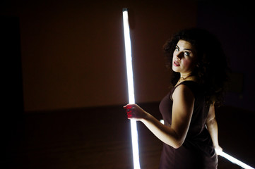Curly brunette plus size model with two long led lamp at black room.