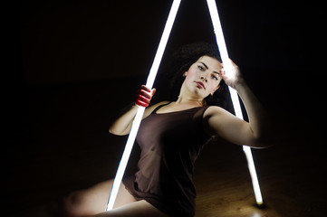 Curly brunette plus size model with two long led lamp at black room.