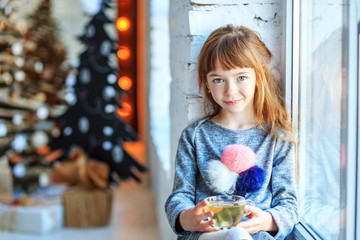 A little child sits on a window sill. The girl is drinking tea. Concept Happy Christmas, New Year, holiday, winter, childhood.