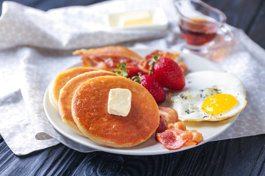 Plate with yummy pancakes, strawberry, fried egg and bacon on wooden table