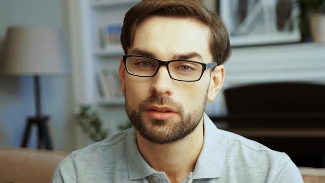 Portrait of young handsome man in glasses looking directly to the camera and smiling sincerely in the cozy living room. Close up
