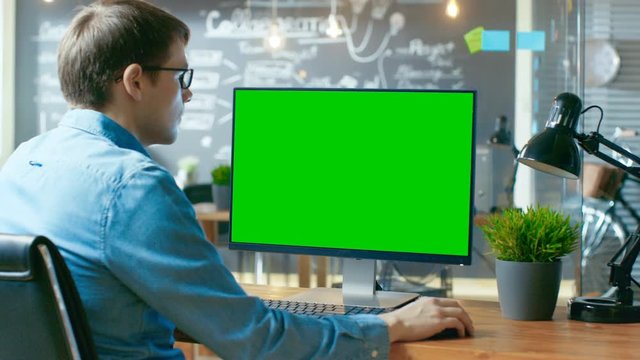 Young Man Works at His Desk on the Personal Computer with Mock-up Green Screen. In the Background His Colleague Works in the Creative Office. RED EPIC-W 8K Camera.