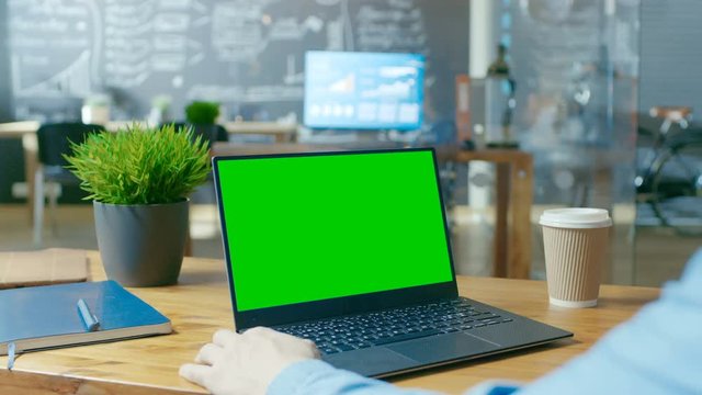 Male Office Worker at Hir Desk Works on a Laptop with Mock-up Green Screen. Over the Shoulder Close-up Footage. In the Background Creative Office. RED EPIC-W 8K Camera.
