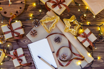 Christmas gift boxes in craft paper wjth satin ribbon, holiday decorations, notebook for planning, lights garland on dark wooden rustic table. Christmas planning concept. Selective focus.