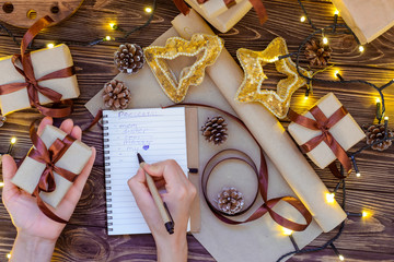 Top view female hands writing gift list in the notebook for a loved people for christmas on wooden table with decor, lights garland, notebook, craft paper. Selective focus. Space for text