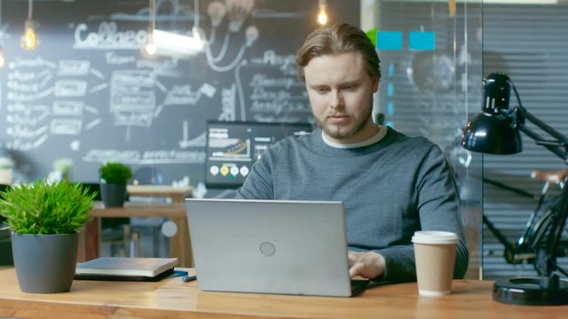 Handsome Young Office Employee Thinks on a Problem Solution While Typing on a Laptop Computer. He's Working in the Creative Stylish Office.